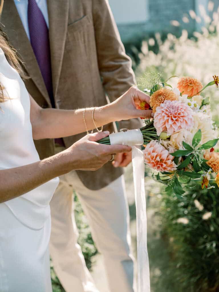 A bride in a white dress adjusts a white ribbon around a vibrant Nantucket wedding bouquet of orange and pink flowers, while the groom in a brown jacket and purple tie stands beside her outdoors.