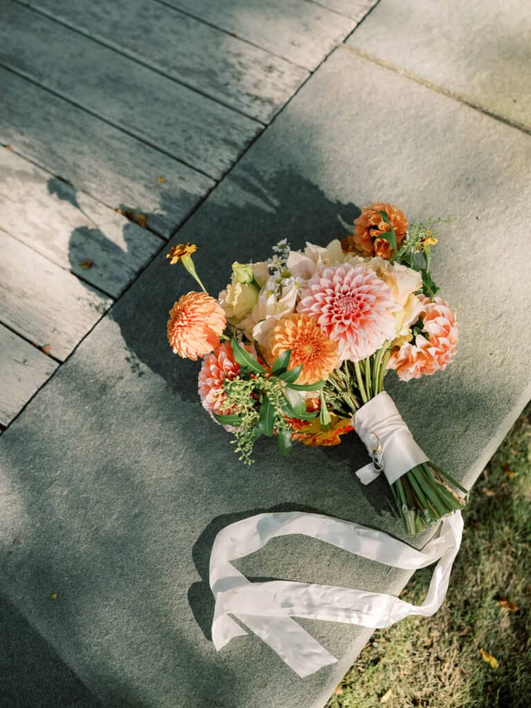 A nantucket bridal bouquet of orange and pink dahlias, yellow flowers, and greenery with a long white ribbon rests on a stone surface, bathed in sunlight beside a wooden pathway.