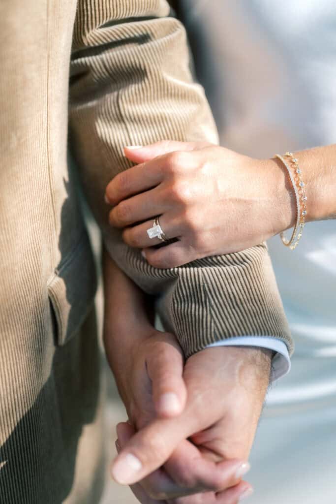 A close-up of a Nantucket bride and groom holding hands—one in a brown corduroy jacket, the other with a white sleeve, gold bracelet, and engagement ring sparkling on their finger.