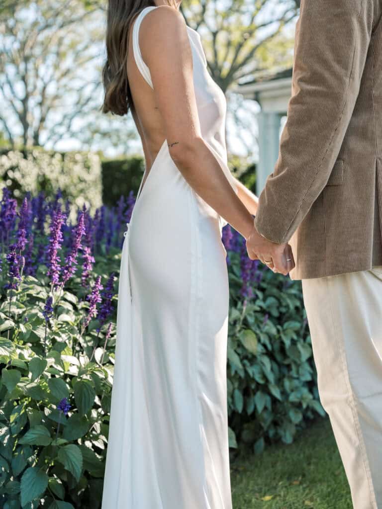 A nantucket bride and groom stand holding hands outdoors; the woman wears a white sleeveless gown and the man wears a tan jacket and cream pants. Purple flowers and greenery surround them under bright daylight.