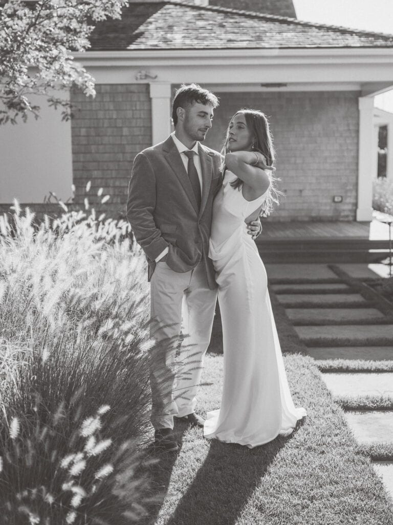 A nantucket wedding couple stands together outdoors in formal attire. The man wears a suit and the woman wears a long dress as they pose closely on a sunlit lawn in front of a building.