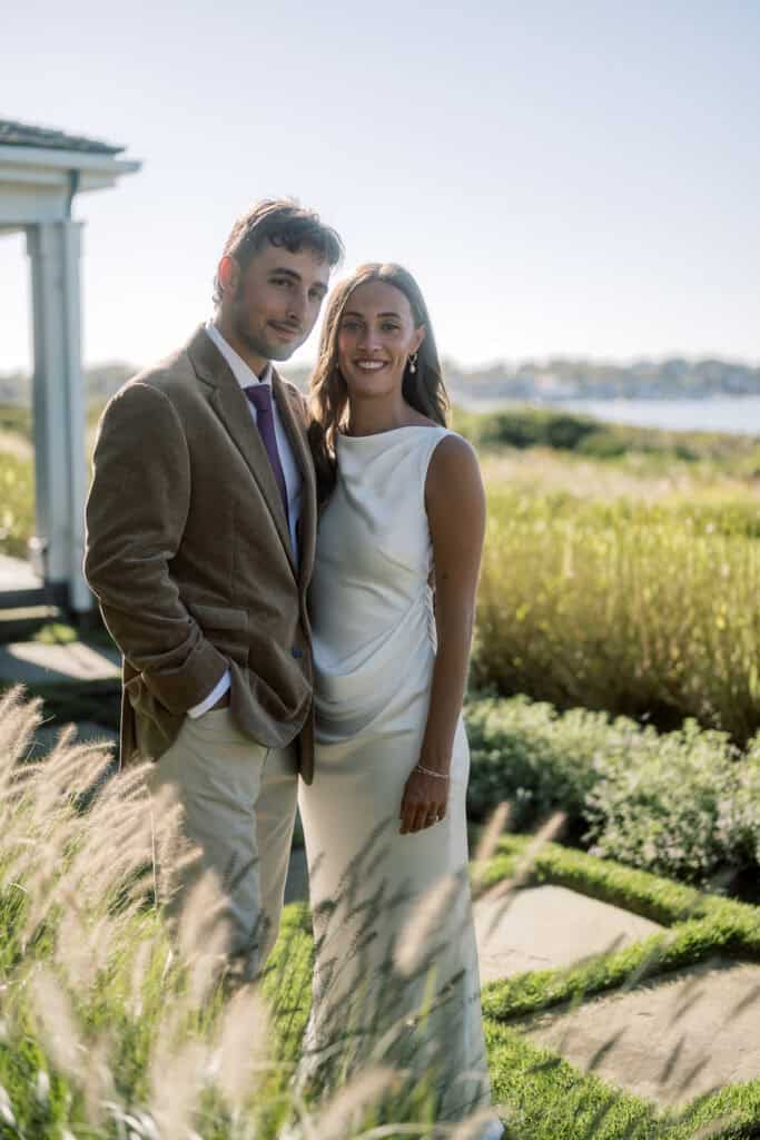 A bride and groom smile outdoors near tall grass, with a building and water behind them under clear skies—capturing a lovely Nantucket wedding portrait. The man wears a brown blazer and tie, while the woman is in a sleeveless white dress.