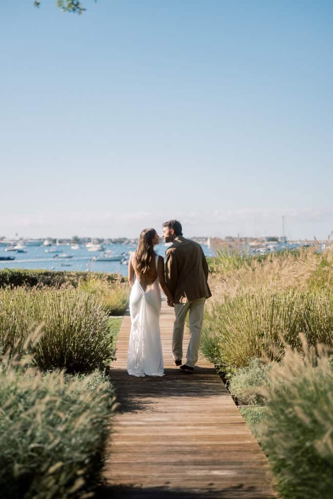 A couple walks on a wooden path through tall grass, the woman in a white dress and the man in a suit, heading toward water and boats under a clear blue sky—a classic Nantucket wedding moment.