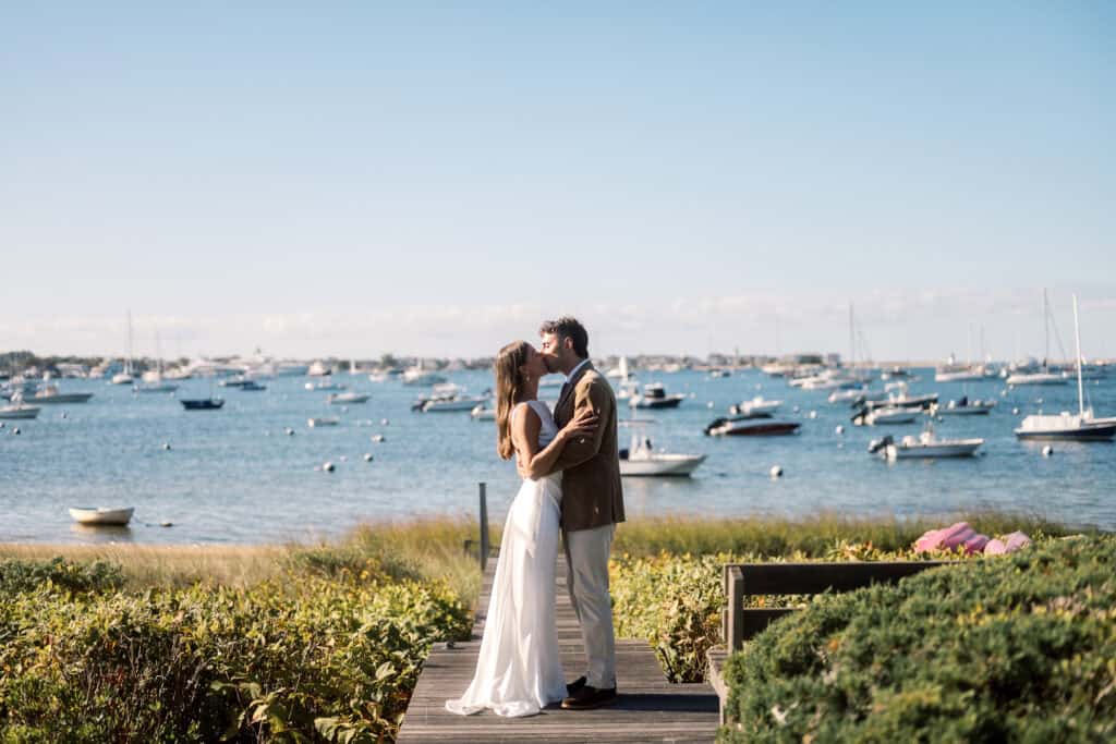 A Nantucket bride and groom share a kiss on a wooden path by the seaside, with boats floating on the water and a clear blue sky in the background.