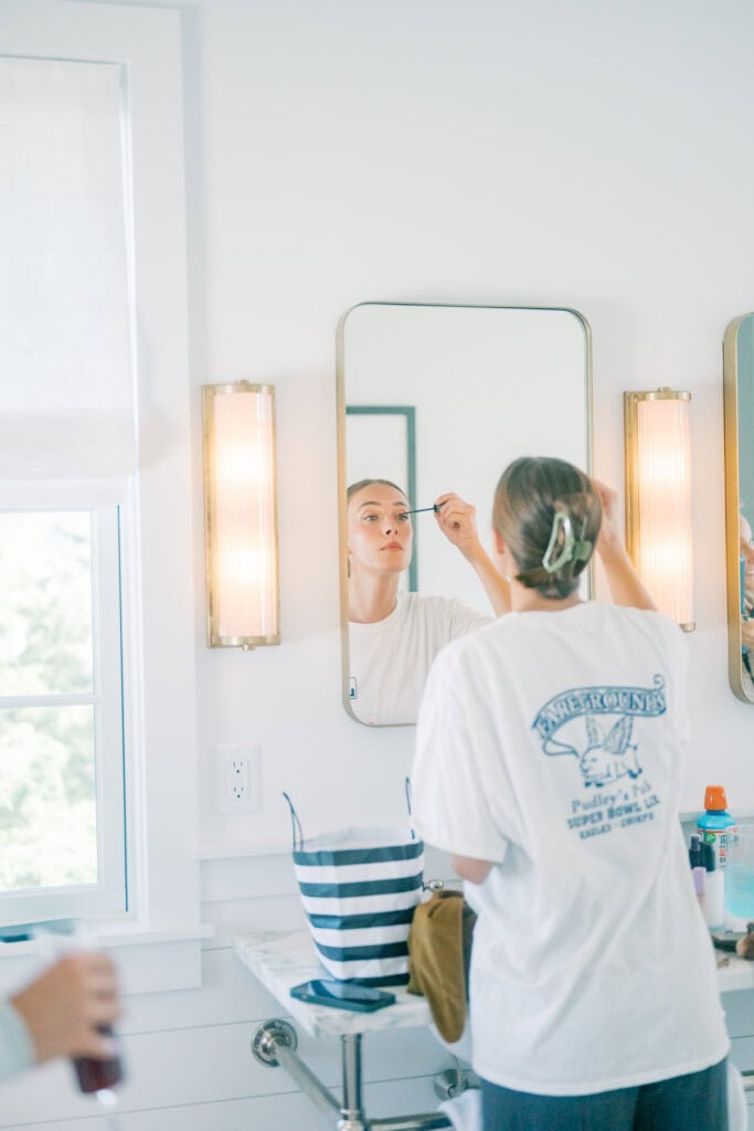 A Nantucket bride stands in front of a bathroom mirror applying makeup, wearing a white t-shirt with graphics on the back. The bright bathroom features white walls, a striped bag, and toiletries on the counter.