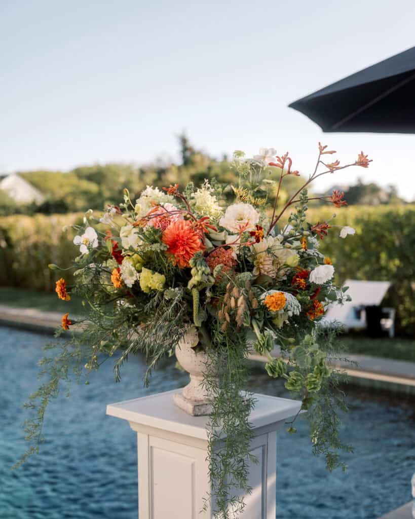 A large, colorful floral arrangement in a white vase sits on a pedestal beside a pool—these vibrant Nantucket wedding florals feature orange, white, and yellow blooms with lush greenery, set outdoors on a sunny day.