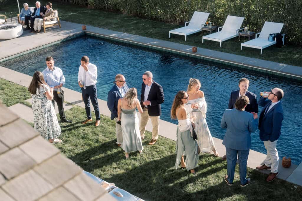 A group of people in semi-formal attire stand and talk by a narrow backyard pool on a sunny day, at what appears to be a relaxed Nantucket wedding, with some guests seated in the background and empty lounge chairs nearby.