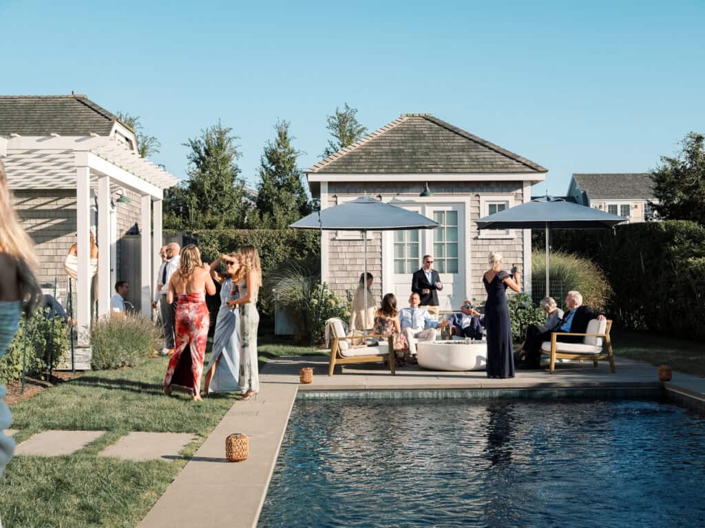 People gather and socialize by a pool in a sunny backyard, reminiscent of a relaxed Nantucket wedding, with some seated under umbrellas and others chatting. Greenery and small houses provide a charming backdrop to the scene.