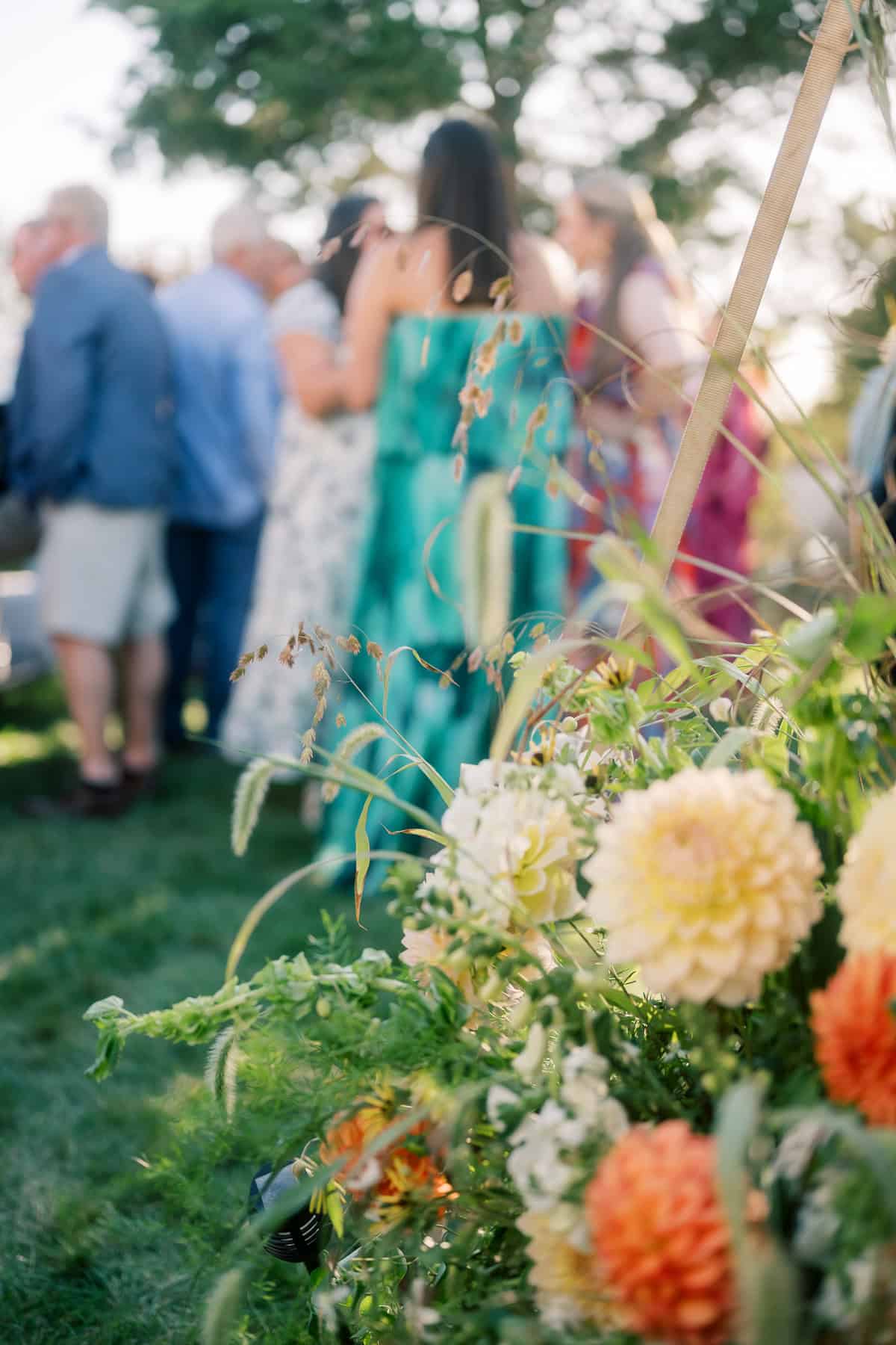 A close-up of yellow and orange flowers with greenery in the foreground; in the blurred background, people in colorful clothing gather outdoors on a sunny day at a Nantucket wedding.