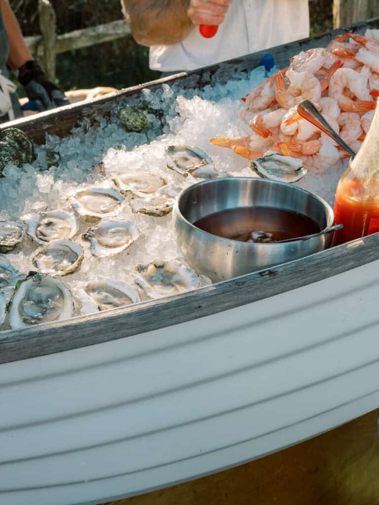 Oysters on the half shell and peeled shrimp are displayed on ice inside a white boat-shaped container, creating an elegant raw bar perfect for a Nantucket wedding, with cocktail sauce and hot sauce served alongside.