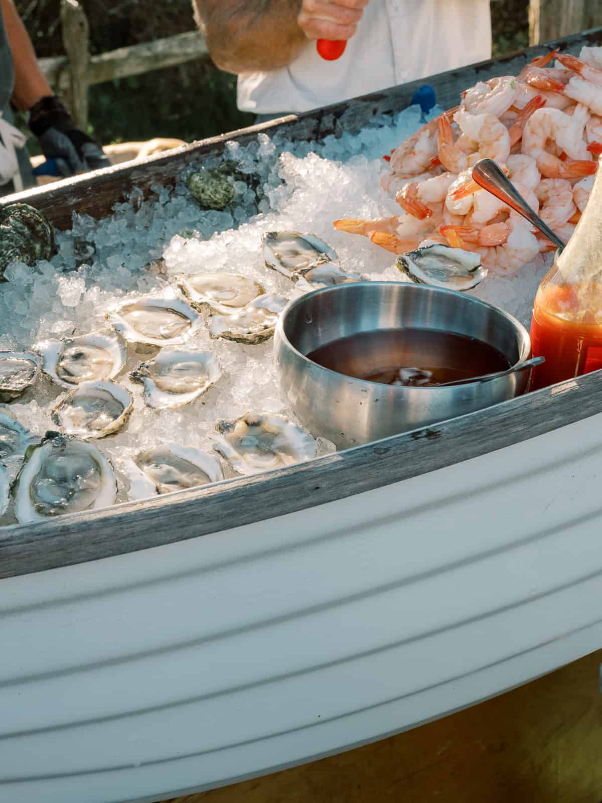 Oysters on the half shell and peeled shrimp are displayed on ice inside a white boat-shaped container, creating an elegant raw bar perfect for a Nantucket wedding, with cocktail sauce and hot sauce served alongside.