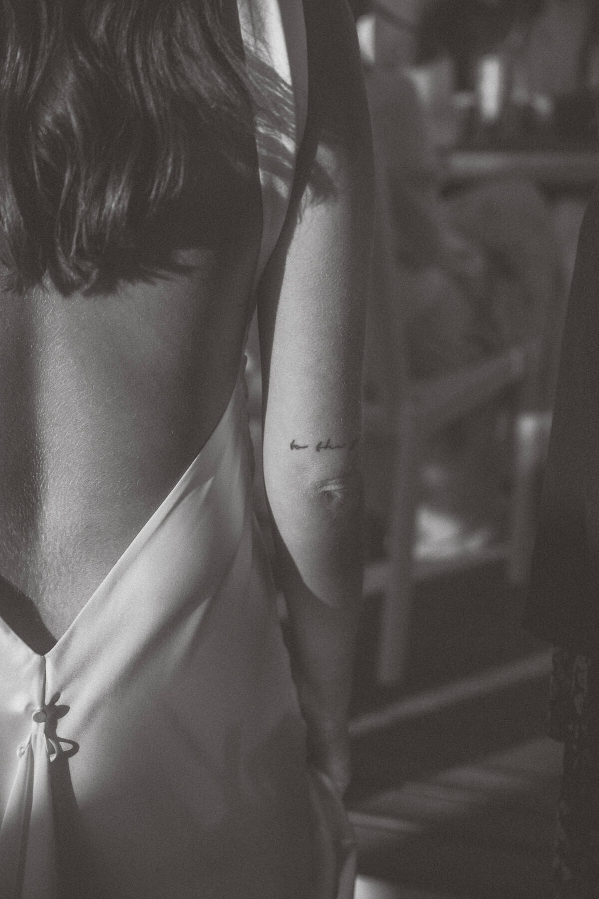 A nantucket bride in a backless dress stands with her back to the camera, revealing a delicate script tattoo on her arm in soft, natural light. The black and white image creates a moody, intimate atmosphere.