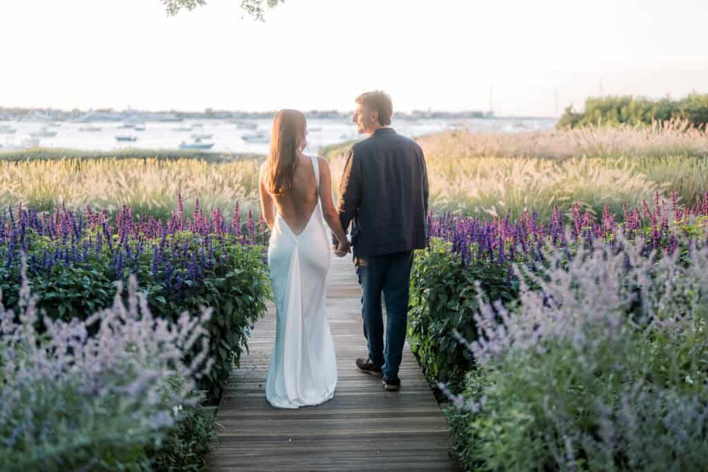 A couple walks hand in hand along a wooden path surrounded by purple flowers, facing the waterfront at sunset. Captured by a Nantucket wedding photographer, the woman wears a white dress, and the man wears a dark jacket and pants.