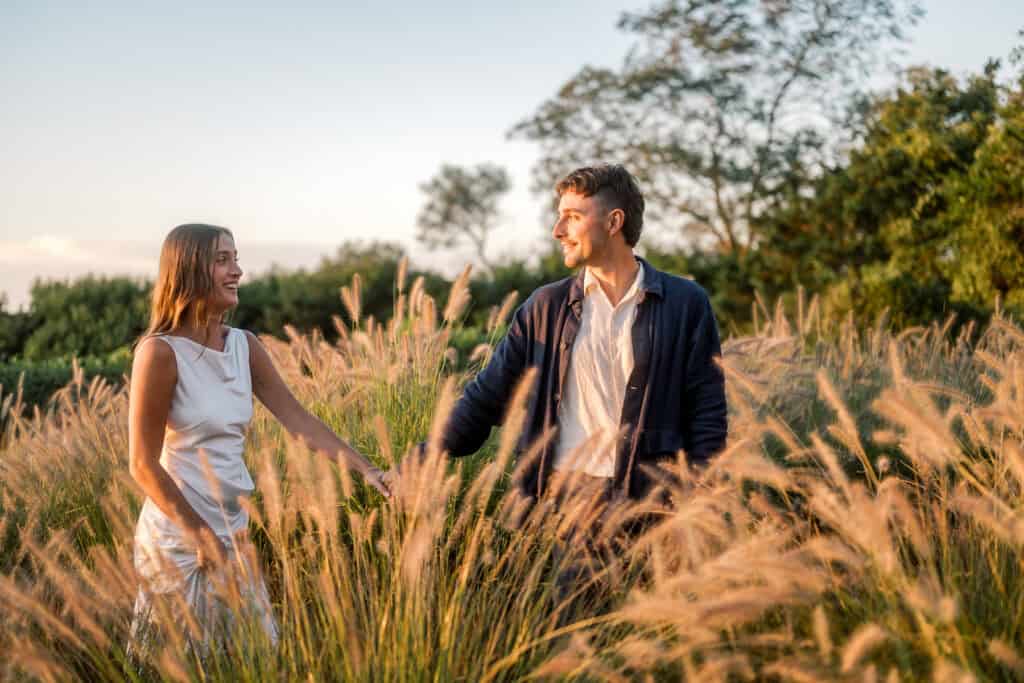 A woman in a white dress and a man in a dark jacket walk hand in hand through tall, golden grass in a sunlit field, smiling at each other with trees and a bright sky—captured beautifully by a Nantucket wedding photographer.