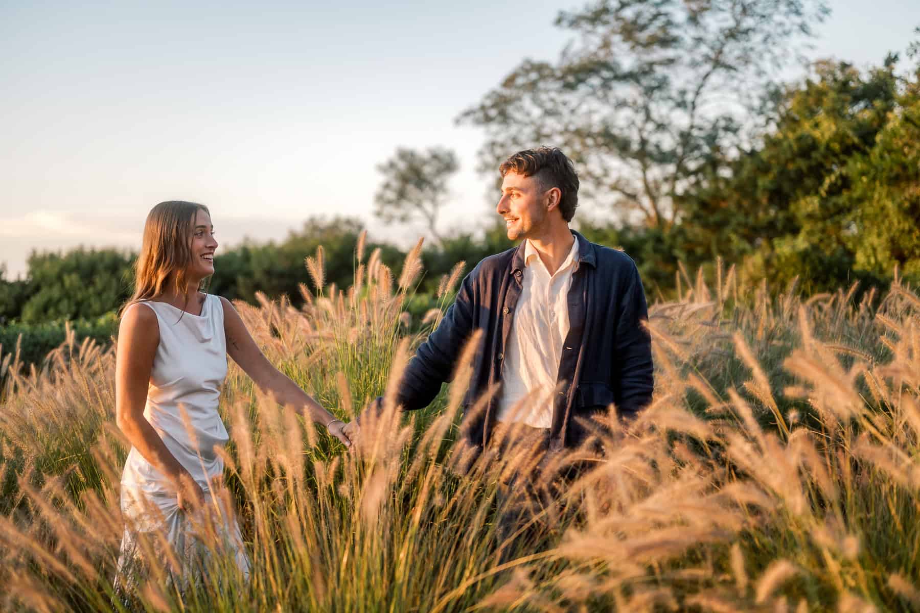 A woman in a white dress and a man in a dark jacket walk hand in hand through tall, golden grass in a sunlit field, smiling at each other with trees and a bright sky—captured beautifully by a Nantucket wedding photographer.