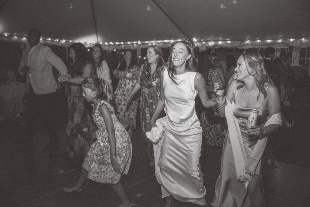 A group of people, including women in dresses and a young girl, dance together under string lights at an indoor event, captured beautifully by a nantucket wedding photographer.