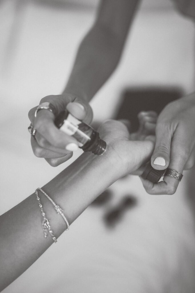 Black and white photo by a Nantucket wedding photographer of a person applying perfume from a small bottle onto the bride's wrist. Both wear rings, one has a delicate bracelet, and the softly blurred background adds to the intimate moment.