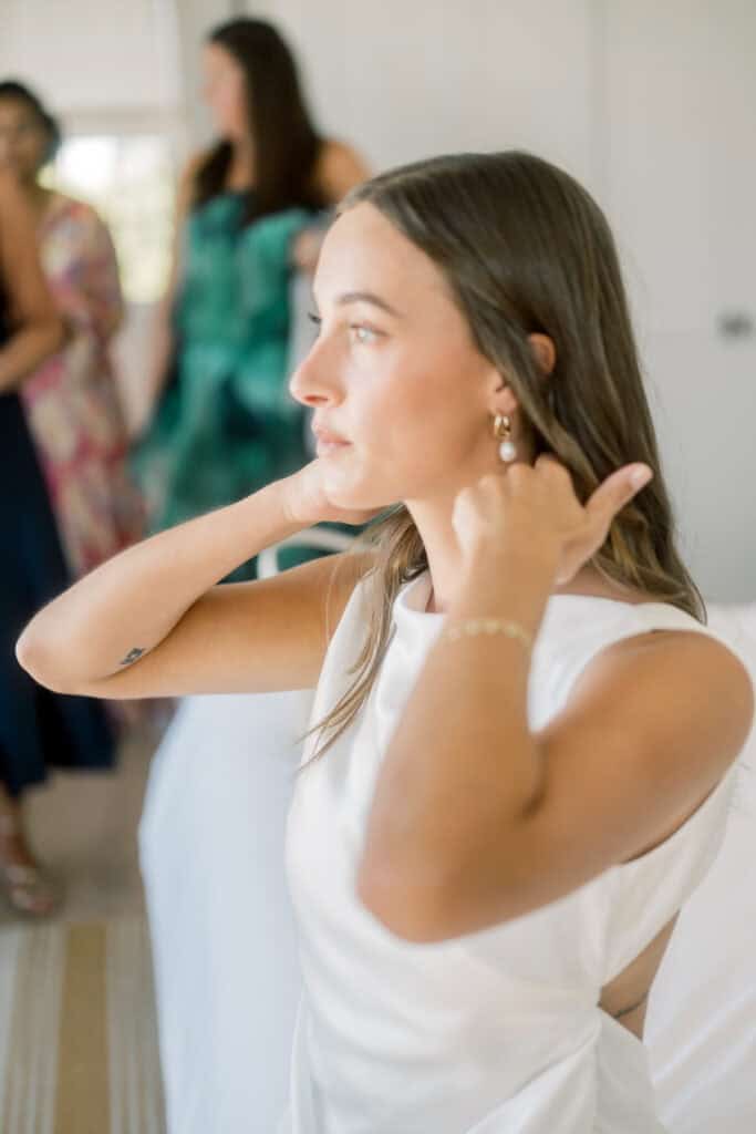 A bride in a white dress adjusts her earring, looking to the side. She has long brown hair and a subtle tattoo on her arm. In the background of this Nantucket wedding, several women in colorful dresses are slightly out of focus.