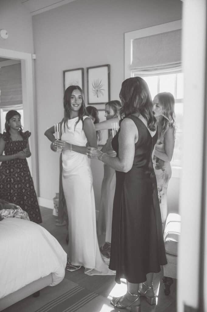 A bride in a white gown and several women stand together in a softly lit room, smiling and holding candles. Captured by a Nantucket wedding photographer, the candid scene feels joyful with natural light streaming through a window.