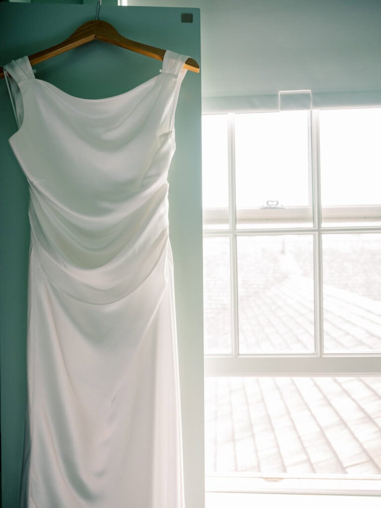 A simple white satin wedding gown hangs on a wooden hanger against a blue wall, with sunlight streaming through a large window nearby—perfect for an intimate Nantucket wedding.