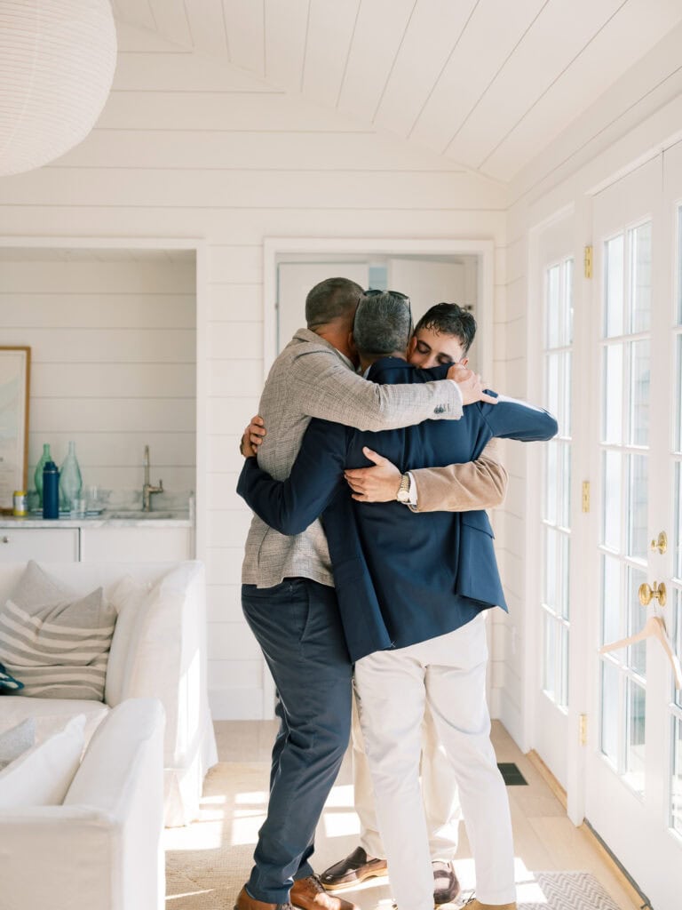 Three men, likely brothers or a groom with his closest friends, stand in a bright, sunlit room, warmly hugging each other. Their dressy casual clothes and the cozy setting evoke the joyful spirit of a Nantucket wedding morning.
