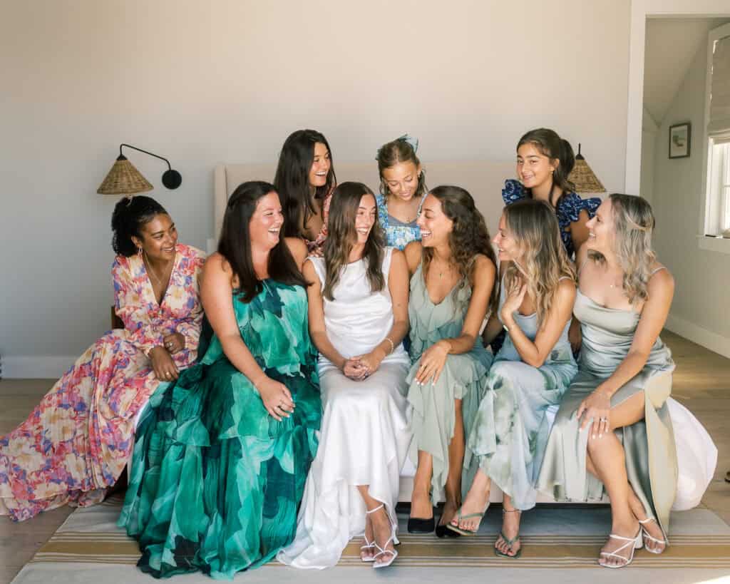 Nine women in colorful dresses sit and stand together indoors, smiling and laughing, with the woman in the center wearing a white dress. This joyful moment captures a bridal party celebrating a nantucket wedding.