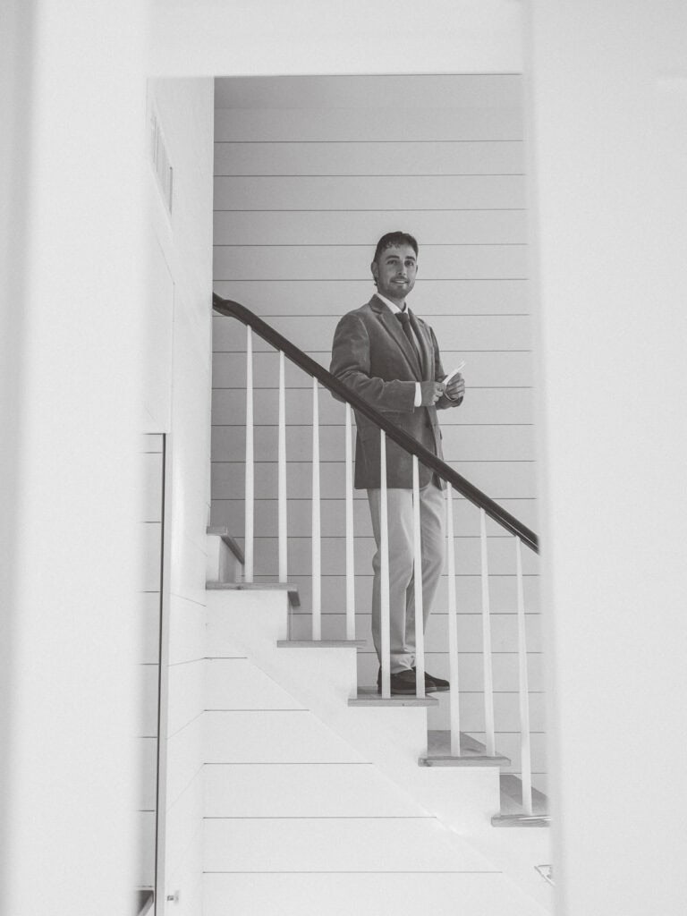 A groom in a suit stands on a staircase, holding something in his hands and looking forward. The setting is modern and minimalist, with white walls and horizontal paneling. The black-and-white image evokes the elegance of a Nantucket wedding.