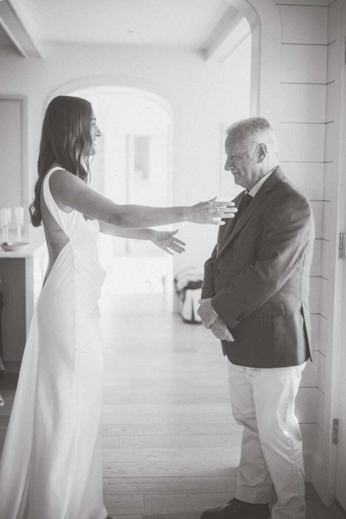 A bride in a white dress with open arms shares a heartfelt first look with her father in a suit and light-colored pants, standing indoors in a bright room during a Nantucket wedding.