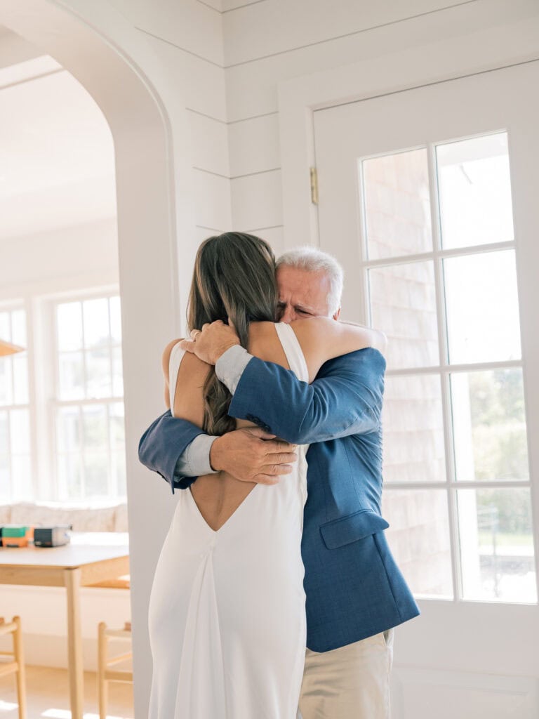 A bride in a white dress hugs her father in a blue suit jacket during an emotional first look inside a bright room with large windows. The embrace appears affectionate and heartfelt.