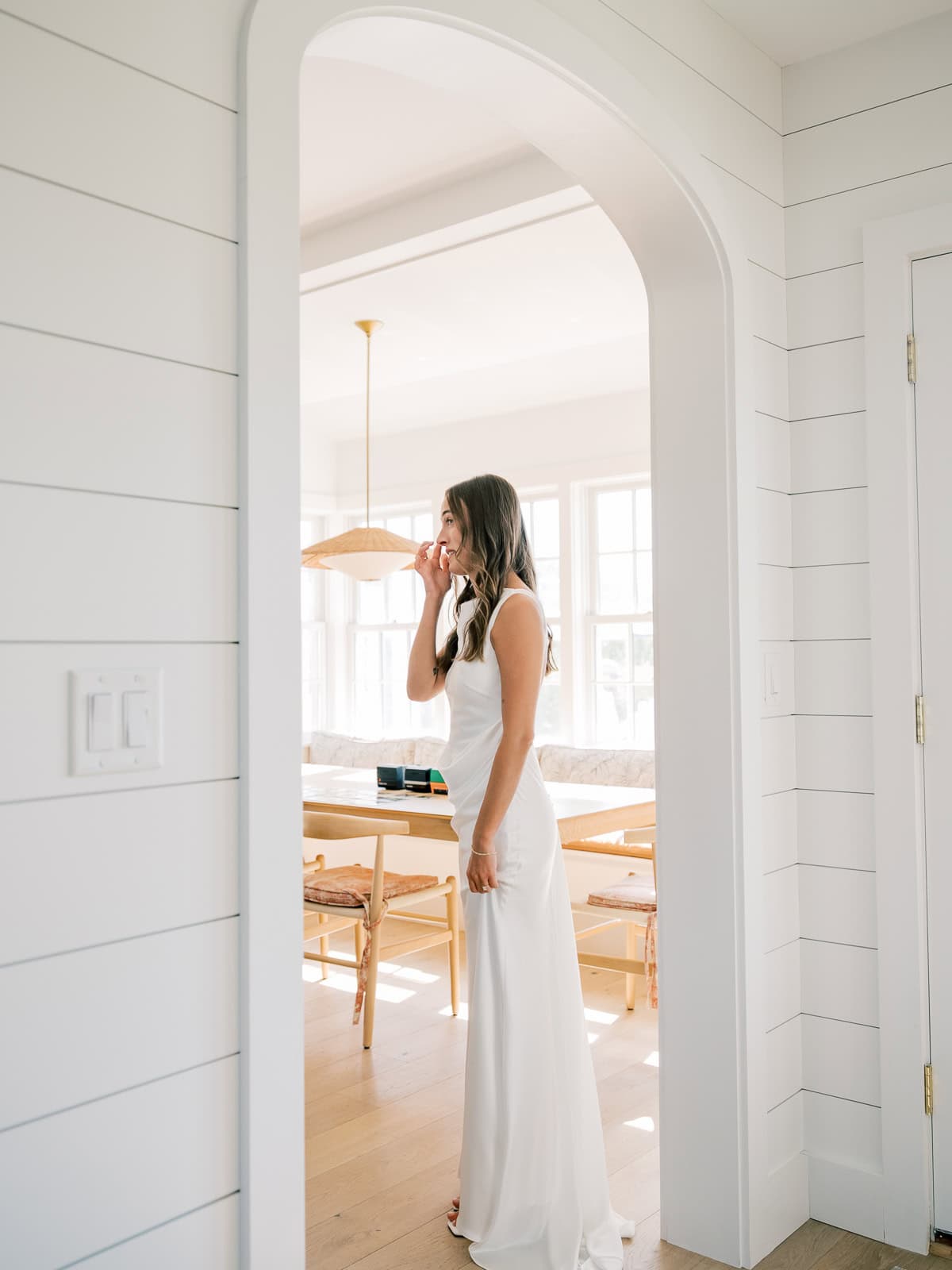 A Nantucket bride in a white dress stands in a bright, modern room with white walls and wooden furniture, talking on the phone—captured beautifully by Nantucket Wedding Photographer Kaity Farrell.