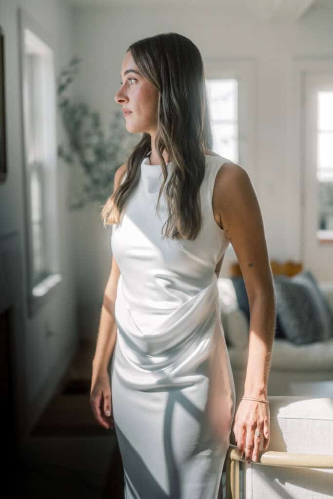 A Nantucket bride with long brown hair stands indoors near a window in a sleeveless white dress. Sunlight casts shadows on her as she gazes outside, and the softly decorated room behind her is bright and inviting.
