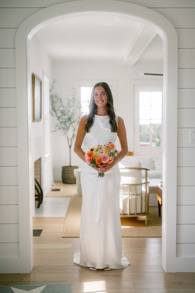 A radiant bride in a simple white gown stands framed by a white archway for her bridal portrait, smiling and holding a bouquet of colorful flowers in a sunlit, cozy room—perfect for an intimate Nantucket wedding.