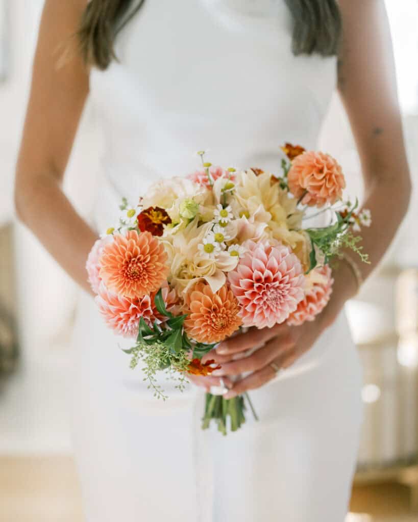 A woman in a white dress holds a stunning bridal bouquet of peach and cream dahlias, small white flowers, and greenery—perfect for a romantic Nantucket wedding. The focus is on the flowers, and her face is not visible.