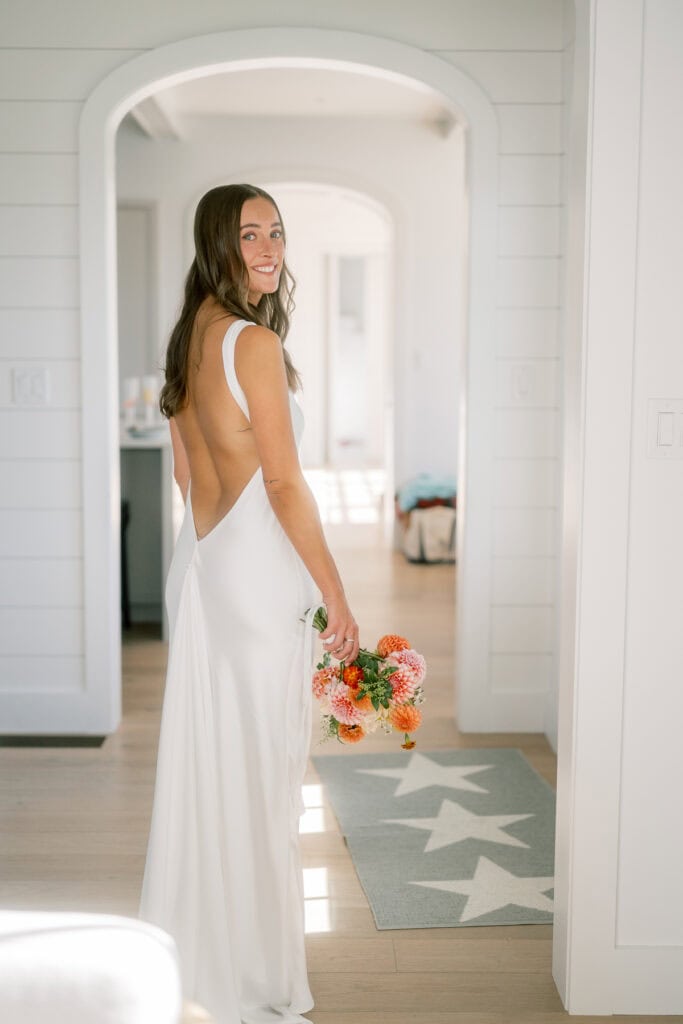 A woman in a backless white dress stands indoors for a radiant bridal portrait, holding a bouquet of colorful flowers and smiling over her shoulder. Sunlight brightens the room, highlighting the rug with white stars on the wooden floor.