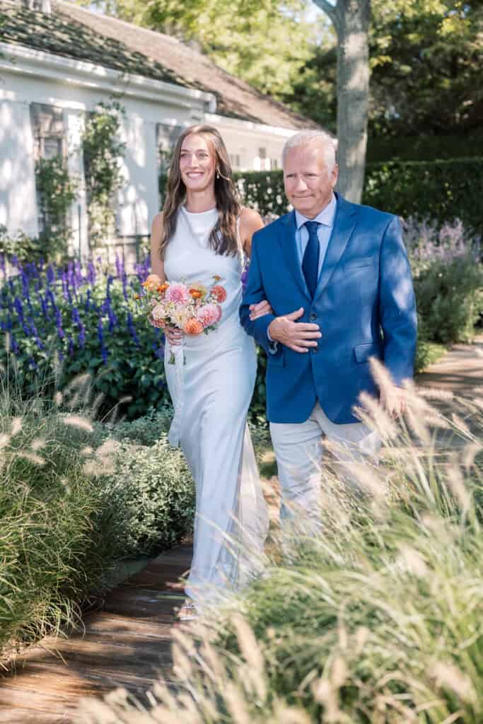 A smiling bride in a white dress holds a bouquet and walks arm-in-arm with her father in a blue jacket along a garden path, surrounded by green plants and purple flowers at a Nantucket wedding ceremony.