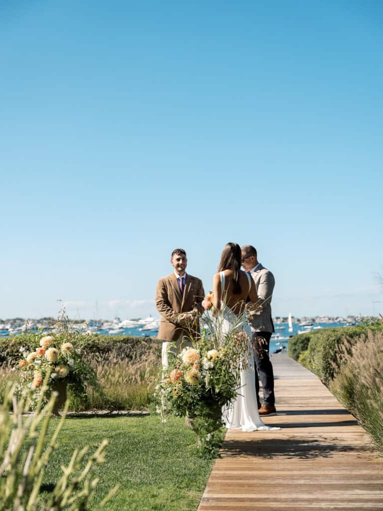 A couple stands on a wooden path by the water, facing an officiant during an outdoor Nantucket wedding ceremony. The scene features clear blue skies, elegant floral arrangements, and boats in the distance.