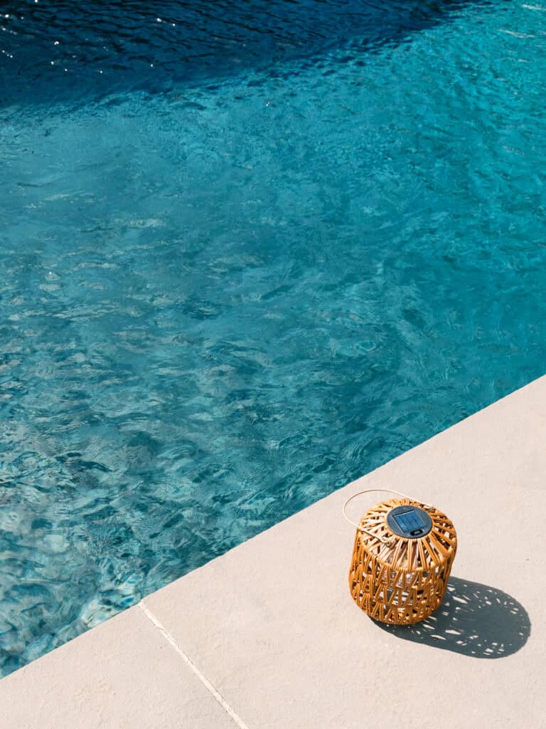 A wicker lantern with a handle sits on the edge of a light-colored pool deck beside clear blue water in a swimming pool, casting a small shadow in the sunlight.