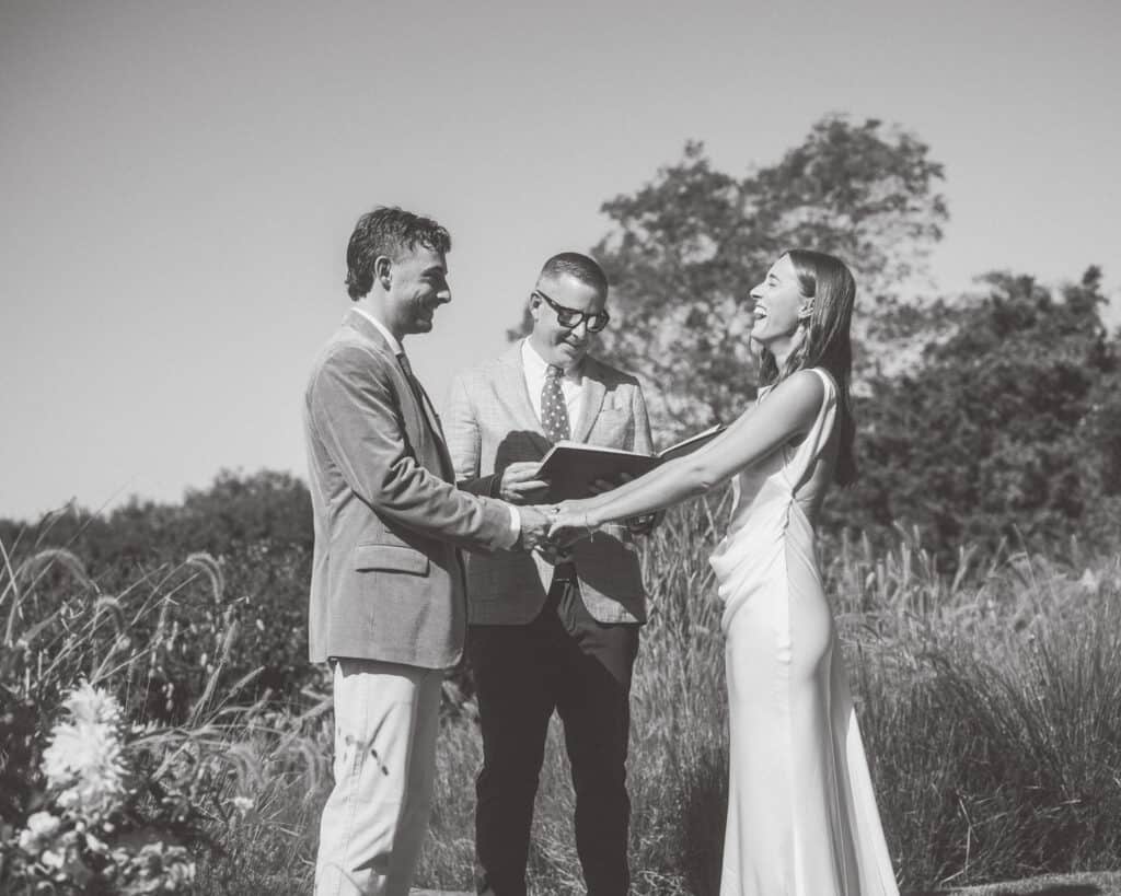 A bride and groom stand holding hands and smiling at each other during a Nantucket wedding ceremony, with an officiant between them and greenery in the background. The photo is in black and white.