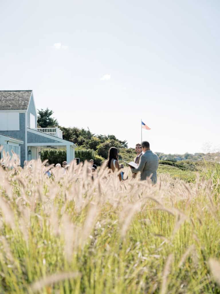 A couple stands together in tall grass near a blue house with an American flag in the background, captured by a Nantucket wedding photographer on a sunny day, surrounded by greenery under a clear sky.