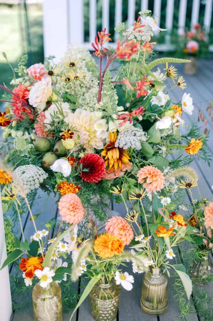 A variety of fresh flowers in shades of orange, yellow, and white arranged in glass vases and a large bouquet on a wooden deck—perfect inspiration for any Nantucket wedding photographer.