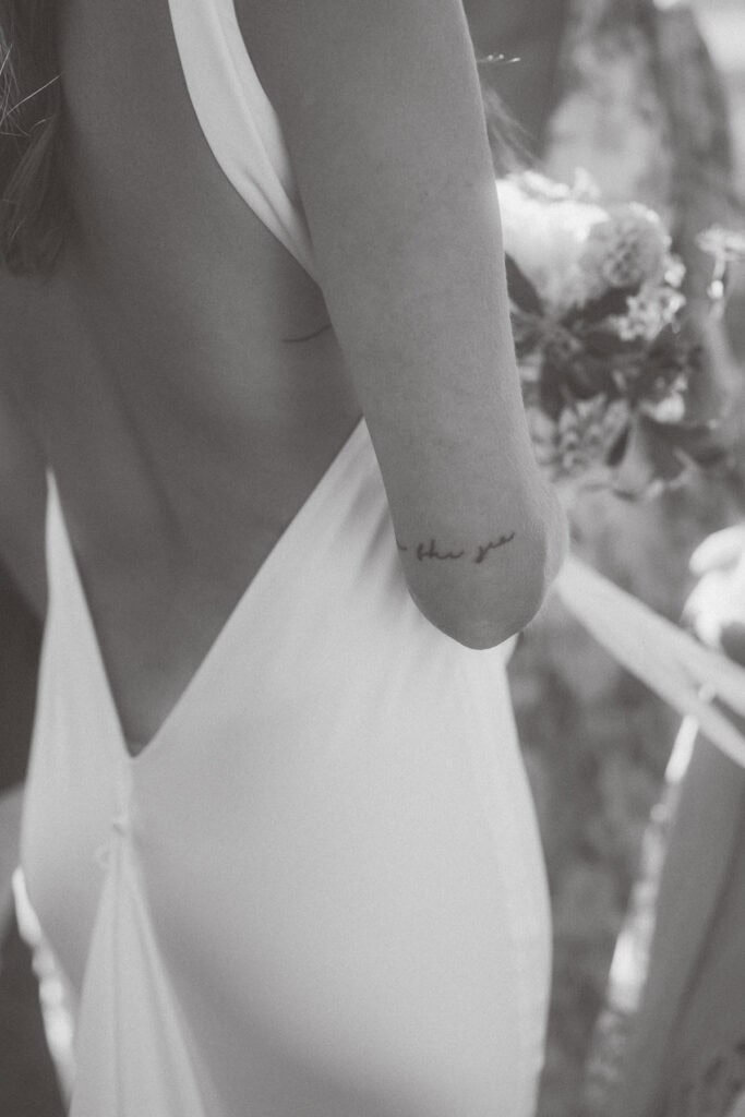 A black-and-white photo shows a nantucket bride in an open-back dress holding a bouquet. A cursive tattoo reading "than you" is visible on her arm, enhancing the image’s soft, romantic, and dreamy atmosphere.