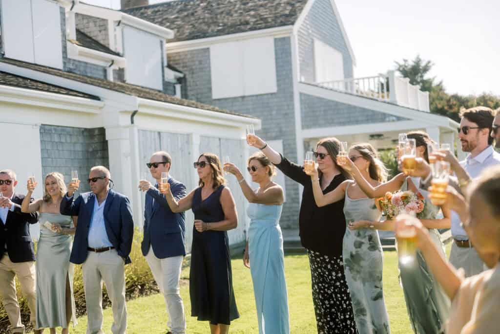 A group of people dressed in semi-formal attire stand outside a gray house, raising glasses in a Nantucket wedding toast on a sunny day, with women in dresses and men in suits.