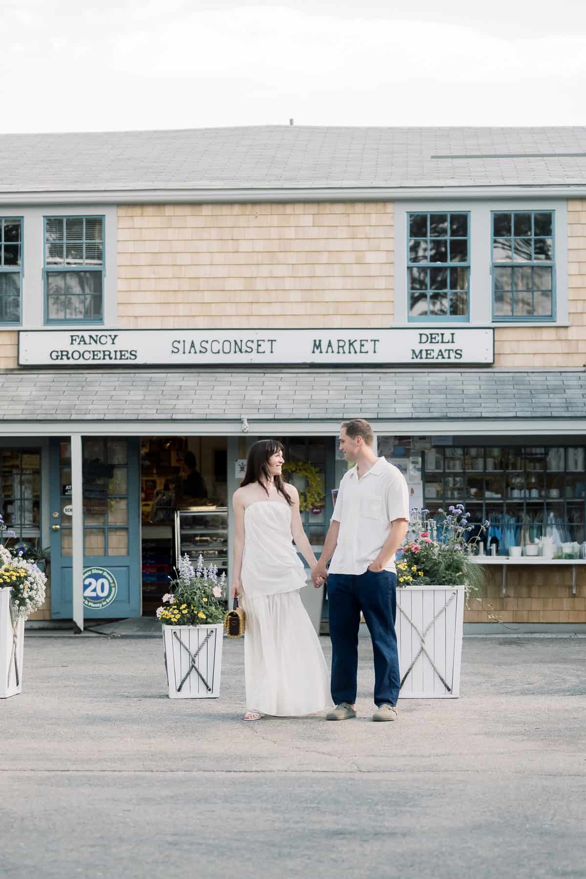 A couple stands holding hands in front of Siasconset Market, a small grocery store with shingle siding and blue trim, captured during their engagement photo session by Nantucket wedding photographer Kaity Farrell.