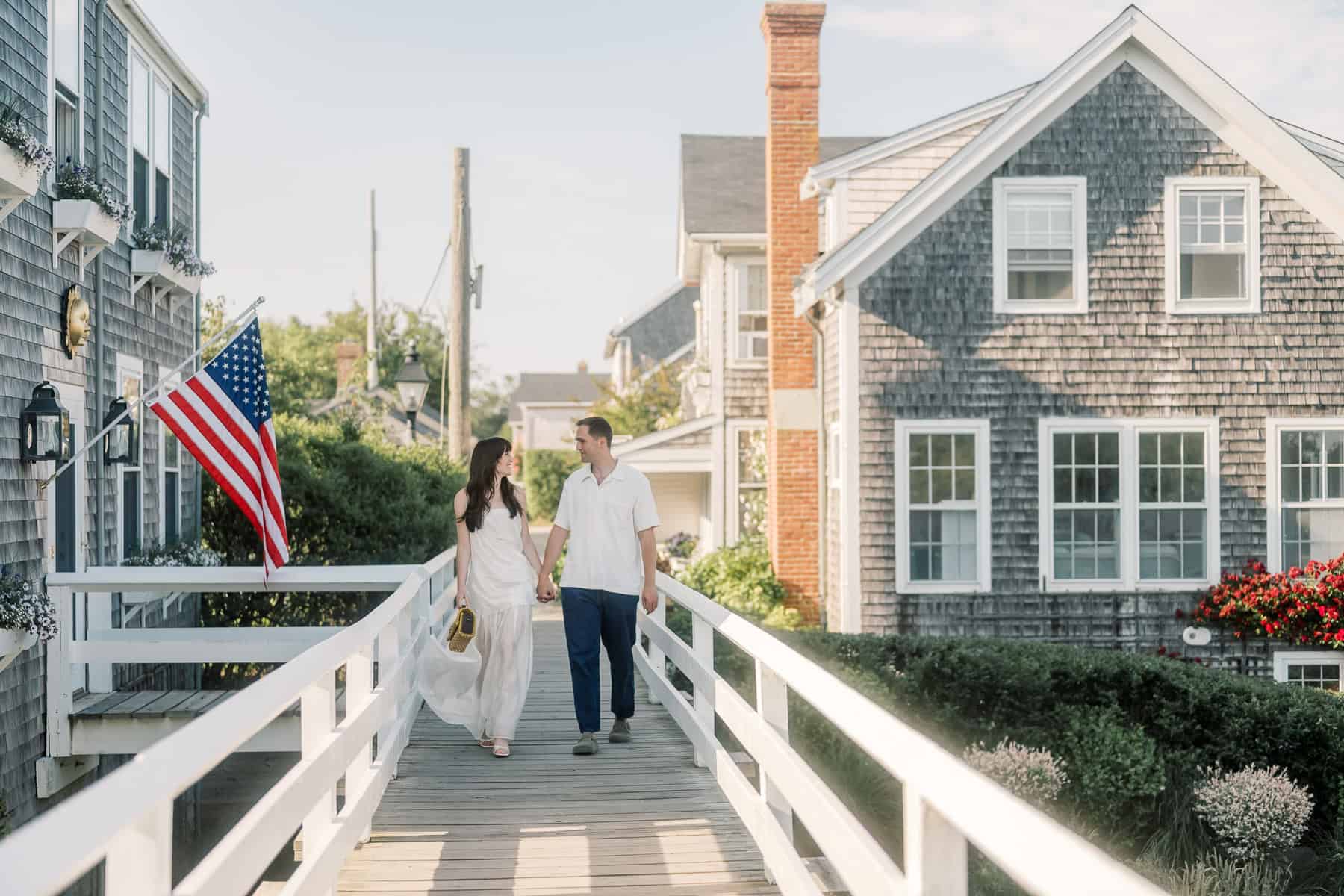 A couple walks hand in hand on the wooden footbridge in Siasconset between two gray shingled houses, with an American flag displayed on the left—captured by Nantucket couples engagement photographer Kaity Farrell.