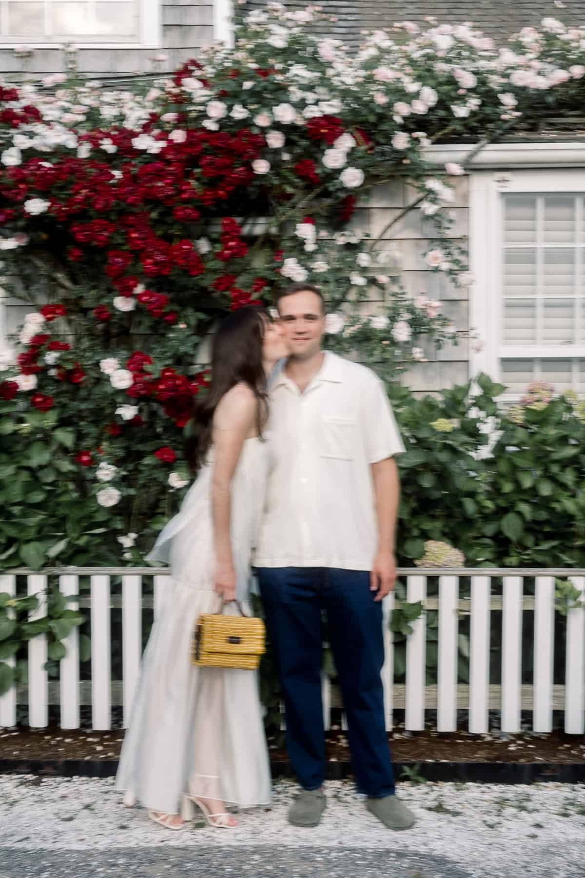 A woman in a white dress kisses a man on the cheek as they stand by a white fence and rose-covered wall, captured beautifully by Nantucket engagement photographer Kaity Farrell.