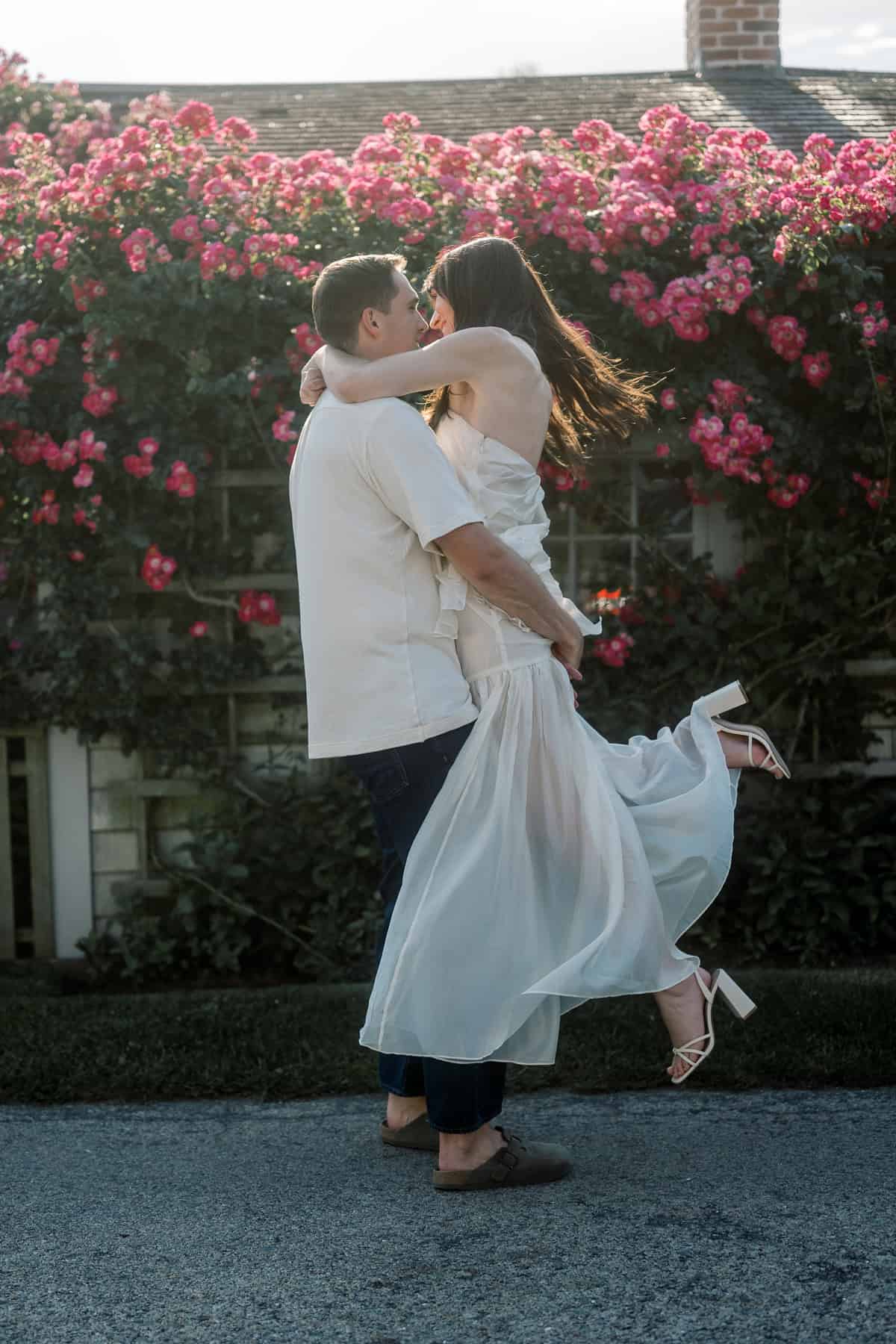 A man lifts a woman in a white dress while they embrace in front of a wall covered with blooming pink flowers during their engagement session, captured by Nantucket couples engagement photographer Kaity Farrell.