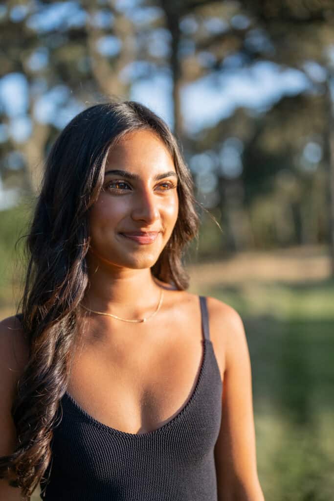 A woman with long dark hair wearing a black sleeveless top stands outdoors in sunlight, with trees and greenery blurred in the background—captured during a Nantucket summer senior session by photographer Kaity Farrell.