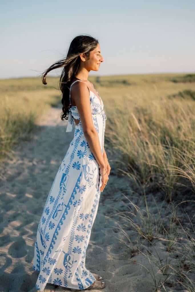 Woman standing on a sandy path in a grassy field, wearing a white and blue summer dress, her hair blowing in the wind under a clear sky—captured by photographer Kaity Farrell for Nantucket beach senior portraits.