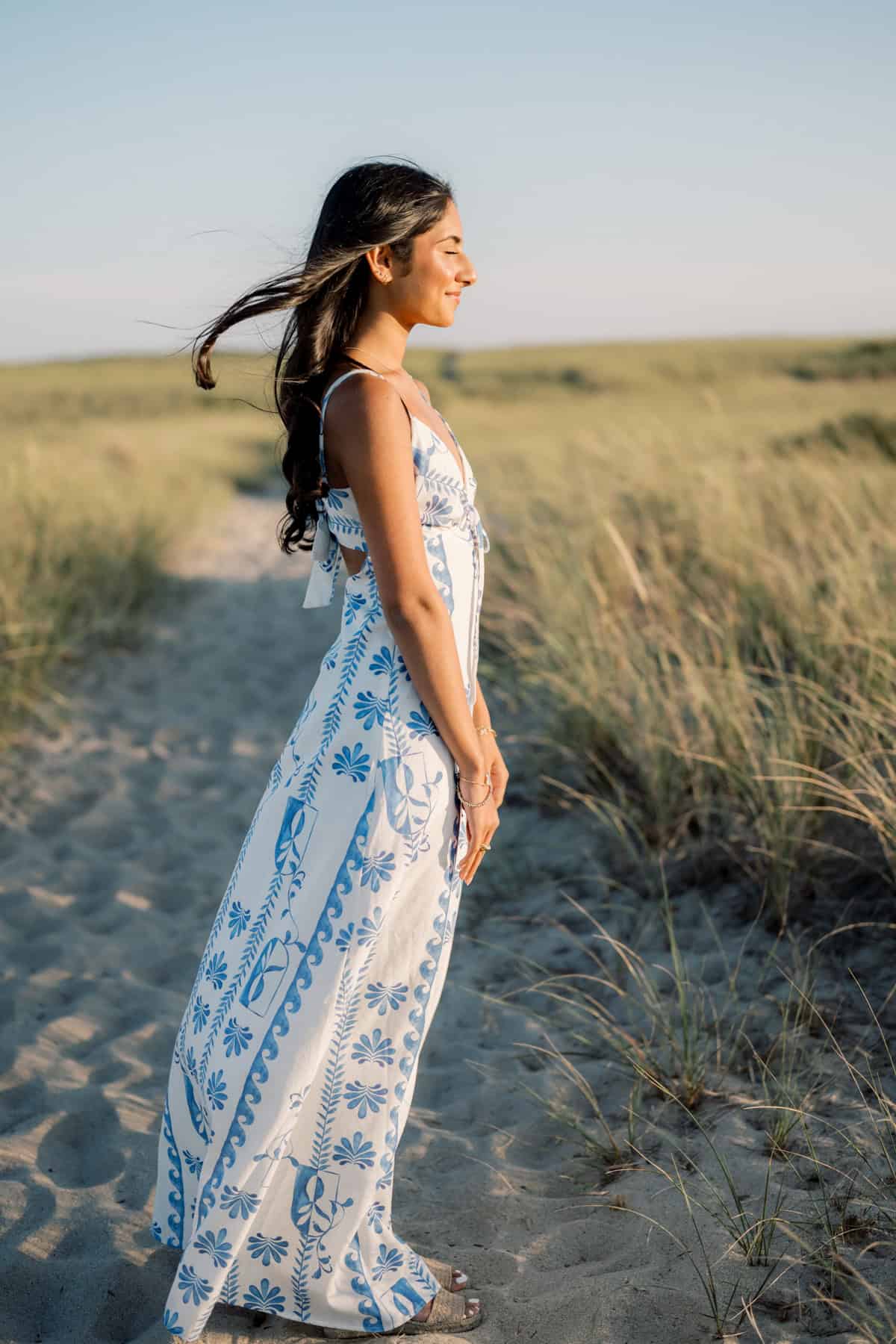 Woman standing on a sandy path in a grassy field, wearing a white and blue summer dress, her hair blowing in the wind under a clear sky—captured by photographer Kaity Farrell for Nantucket beach senior portraits.