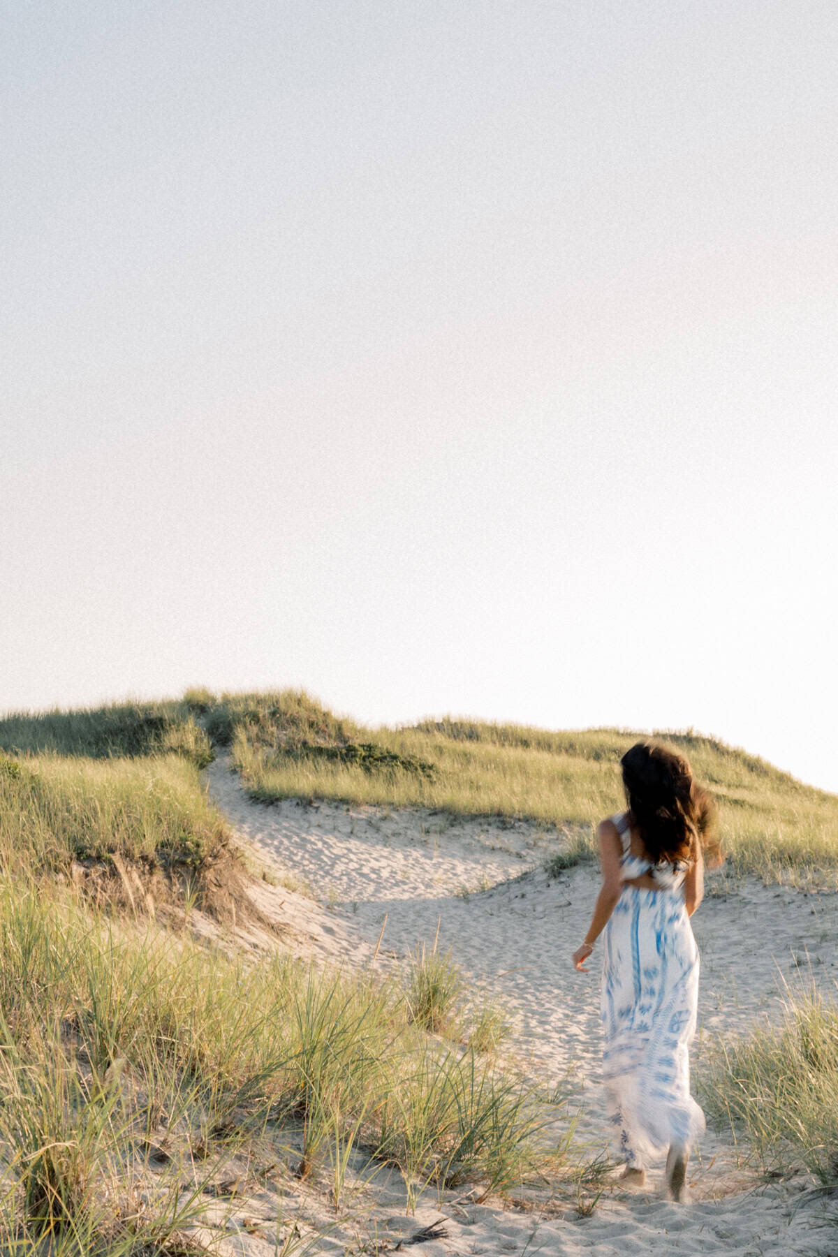 A woman in a white and blue dress walks on a sandy path through grassy dunes under a clear sky, perfect for Nantucket senior portraits by photographer Kaity Farrell.